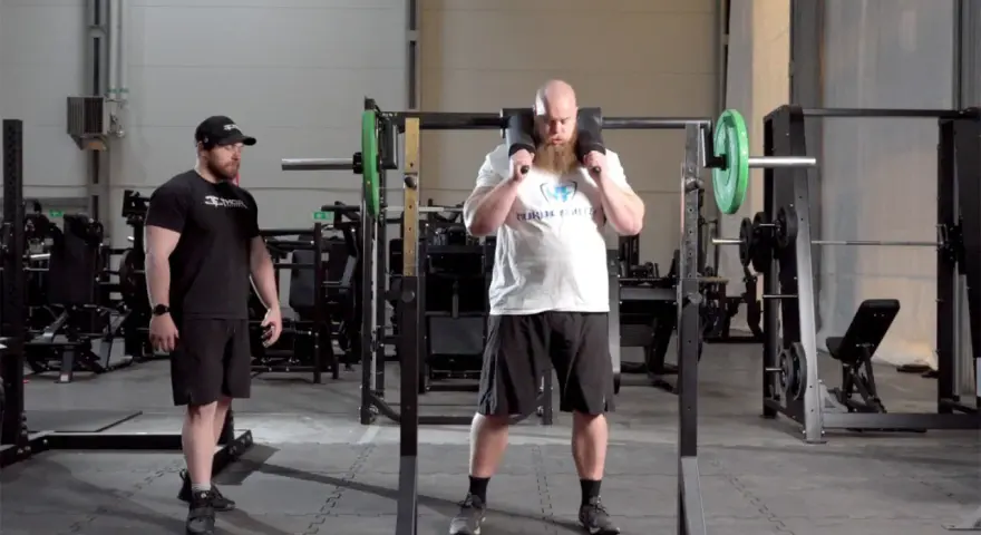 Man med skägg förbereder sig för knäböj med en safety squat bar. En tränare står bredvid.