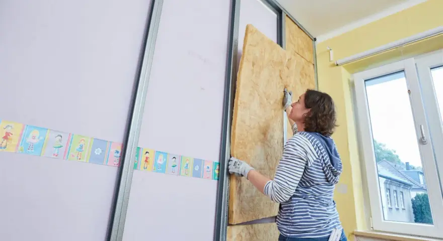 Frau baut eine Trockenbauwand, setzt Dämmmaterial in Metallständer ein.