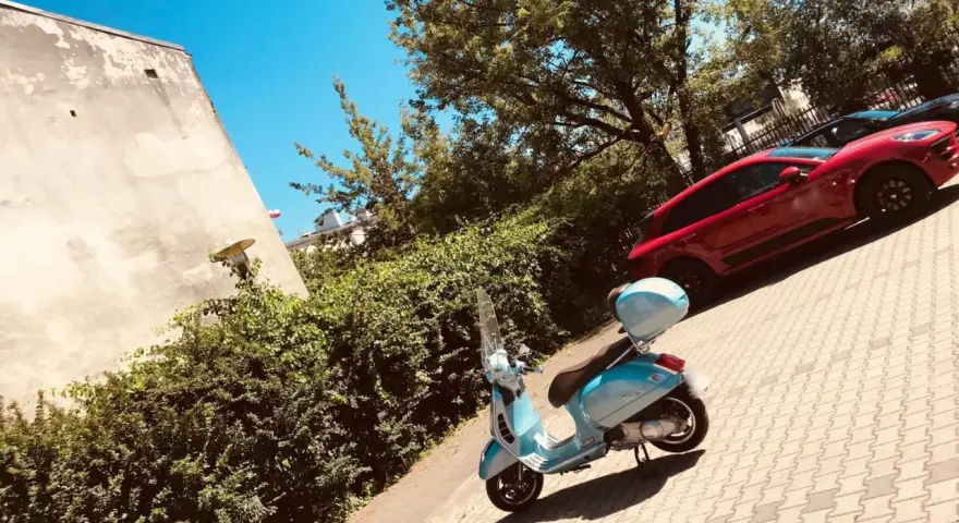 Błękitny skuter czeka na przejażdżkę. W tle czerwony samochód i zielone drzewa. Idealne miejsce na parking moto.