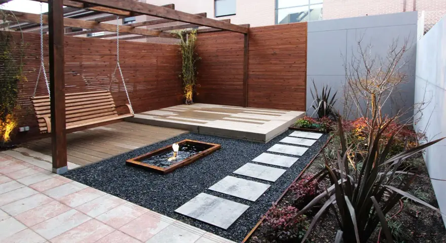 Un columpio de madera cuelga bajo un cenador. Senderos de piedra cruzan grava negra hacia un estanque. Minimalistas jardines modernos.