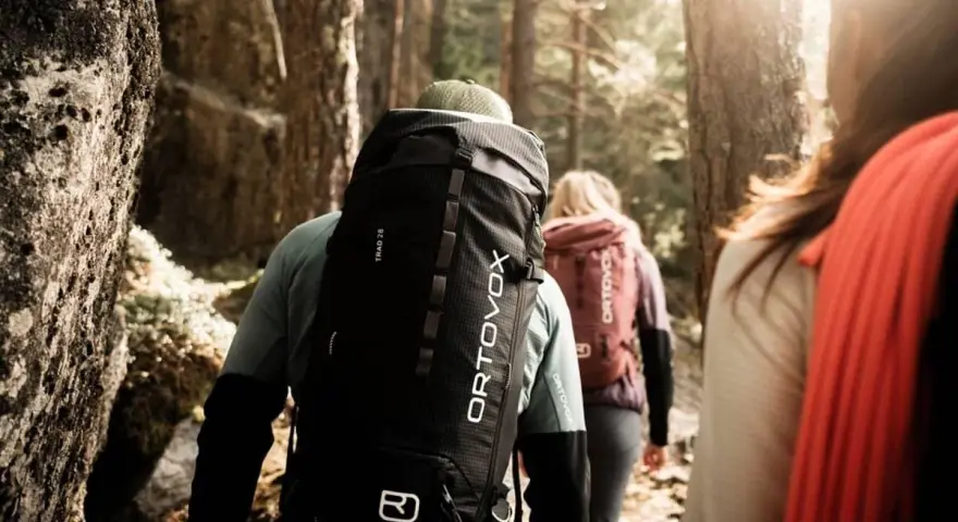 Trois randonneurs avancent dans une forêt ensoleillée, chacun portant un sac à dos Ortovox. Le sac de randonnée noir du premier plan est prêt pour le prochain lavage.