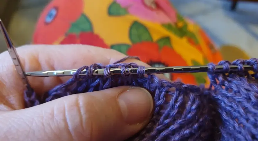 Närbild på en hand som stickar med lila garn. Nålen håller flera maskor, redo för nästa varv i blocka stickning.