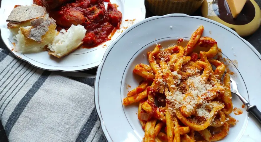 Pyszne maccheroni al ferretto w sosie pomidorowym, posypane parmezanem, obok talerz mięsa w sosie i chleba.