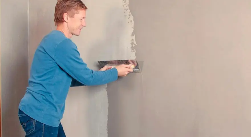 Man med blå tröja och jeans använder en bred spackel för att rolla spackel på en vägg.