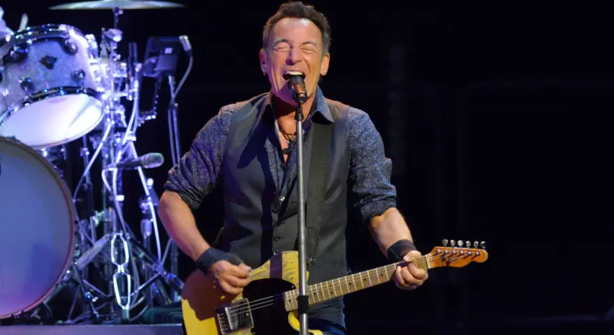 Bruce Springsteen singt und spielt seine ikonische Bruce Springsteen Gitarre auf der Bühne, während die Band im Hintergrund spielt.