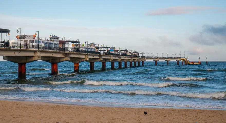 Molo w Międzyzdrojach, jedna z głównych atrakcji, wznosi się nad wzburzonym morzem. Piasek plaży i błękitne niebo tworzą malowniczy krajobraz.