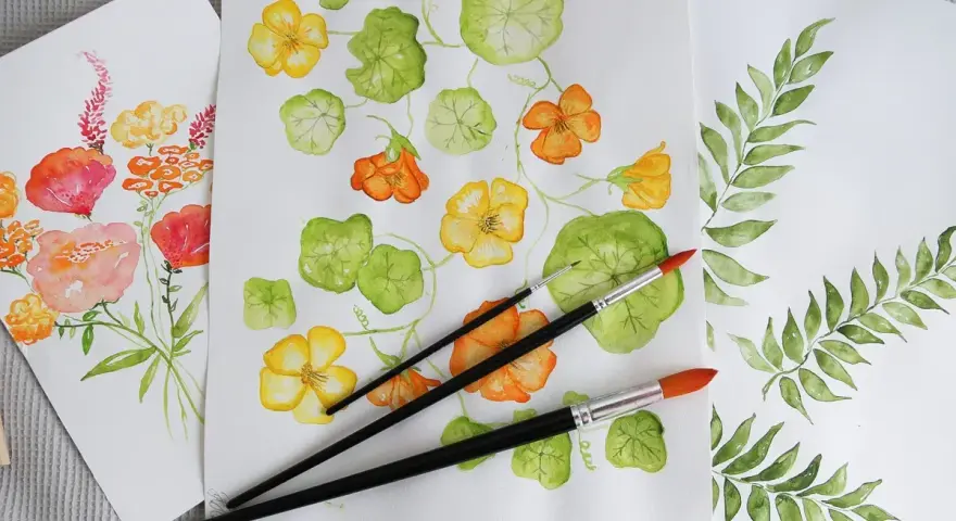 Flera akvarellmålningar med blommor och blad. En målning visar gula och orange blommor med gröna blad, en annan visar en bukett med rosa och orange blommor. Penslar ligger på pappret.