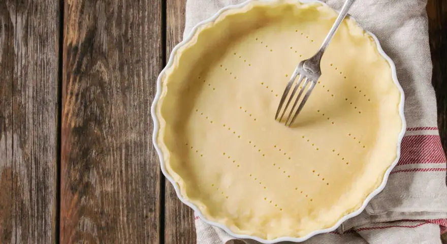 Pâte brisée sans beurre dans un plat à tarte, prête à être garnie. Une fourchette est plantée dans la pâte.