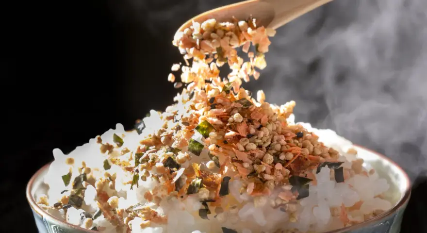 Łyżka sypie na parujący ryż kolorowy furikake. To japońska posypka, która dodaje smaku.