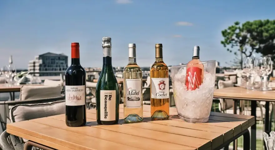 Pięć butelek wina, w tym jedno bordeaux, na drewnianym stole na tarasie z widokiem na miasto.