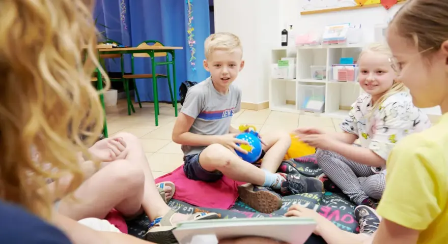 Chłopiec, około 10 lat, siedzi na podłodze z niebieską zabawką. Wokół niego inne dzieci.