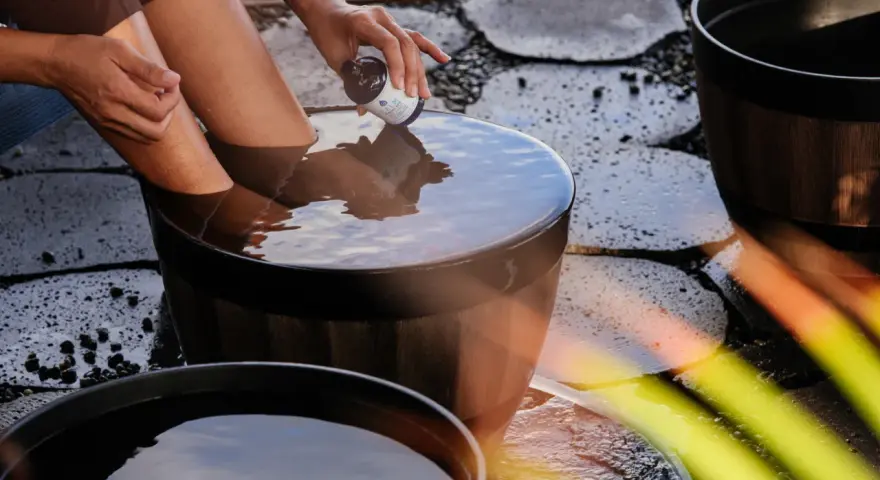 A person adds a few drops from a small bottle to a wooden tub filled with water, creating the best natural foot soak.