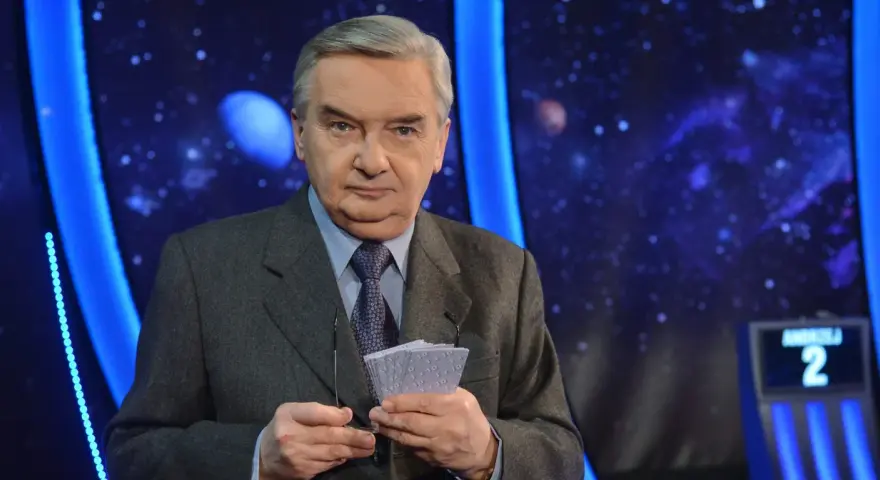 Starsza osoba w garniturze trzyma karty z napisem "1 z 10", gotowa do emisji.