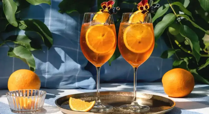 Dwa kieliszki Aperol Spritz z plastrami pomarańczy i kwiatami. Czy aperol można pić sam? Tak, ale najlepiej smakuje w towarzystwie!
