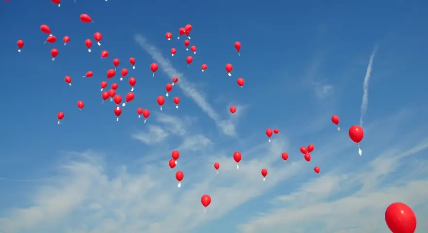 Många röda heliumballonger stiger mot en klarblå himmel med tunna moln.
