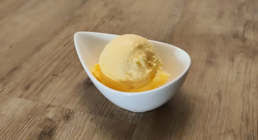 Ein Löffel cremiges Mandarinen Eis in einer weißen Schale auf Holzhintergrund.