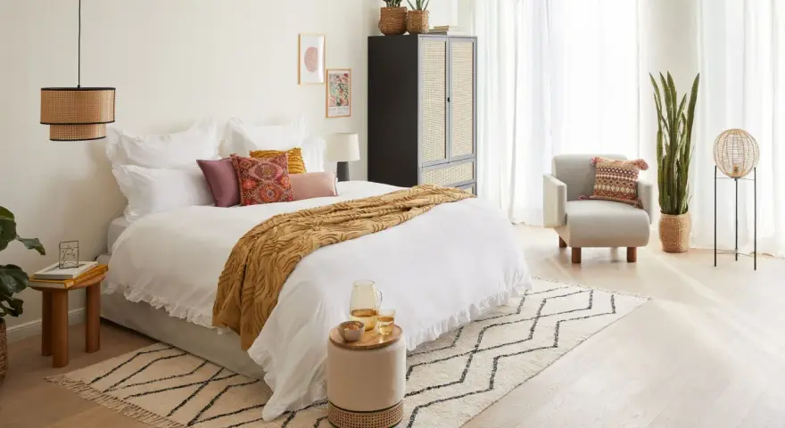 Habitación serena con cama blanca, manta ocre y cojines coloridos. Un armario de ratán y una silla acogedora complementan el estilo feng shui cama.
