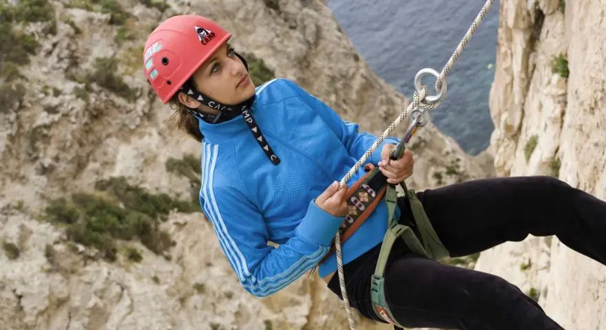 Junge Frau beim Abseilen an einer Felswand, Malta Klettern. Sie trägt einen roten Helm und eine blaue Jacke.