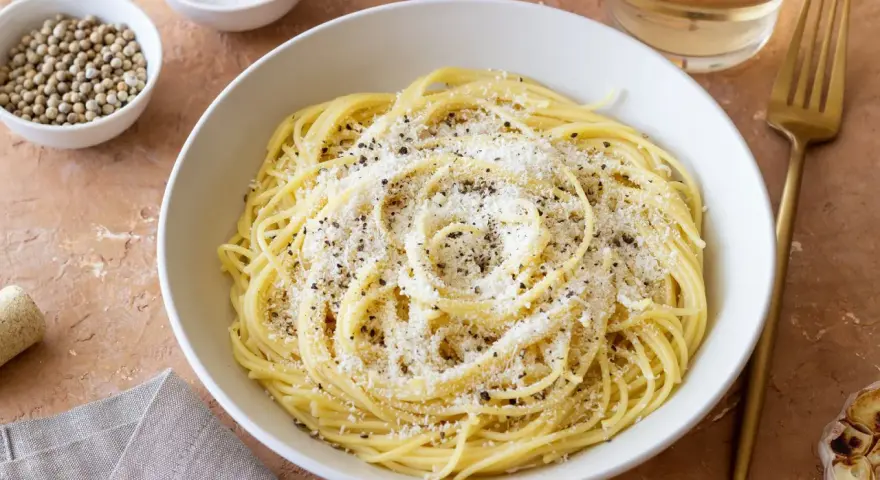 Miseczka spaghetti posypana parmezanem i pieprzem, obok przyprawy i czosnek.