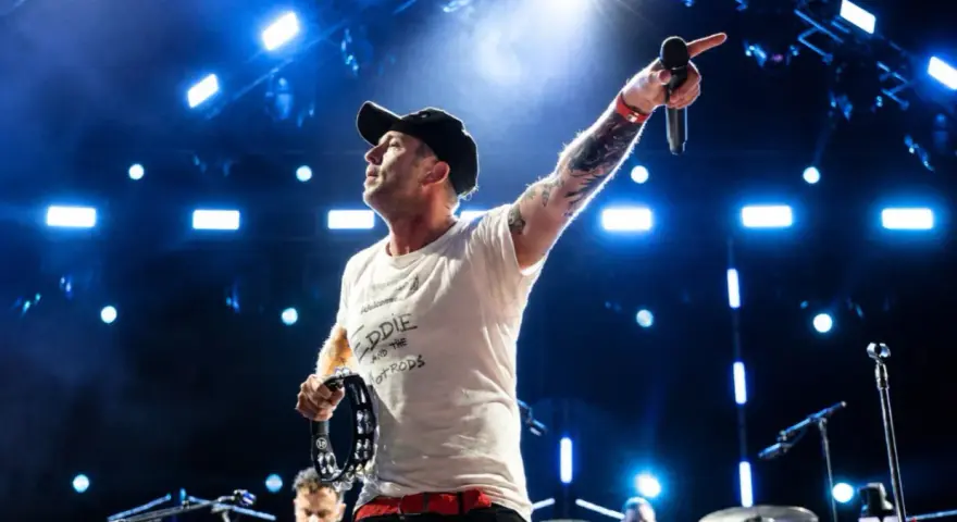 Wokalista z tatuażami i czapką na głowie prowadzi koncert One Republic, wskazując ręką w górę i trzymając tamburyn.