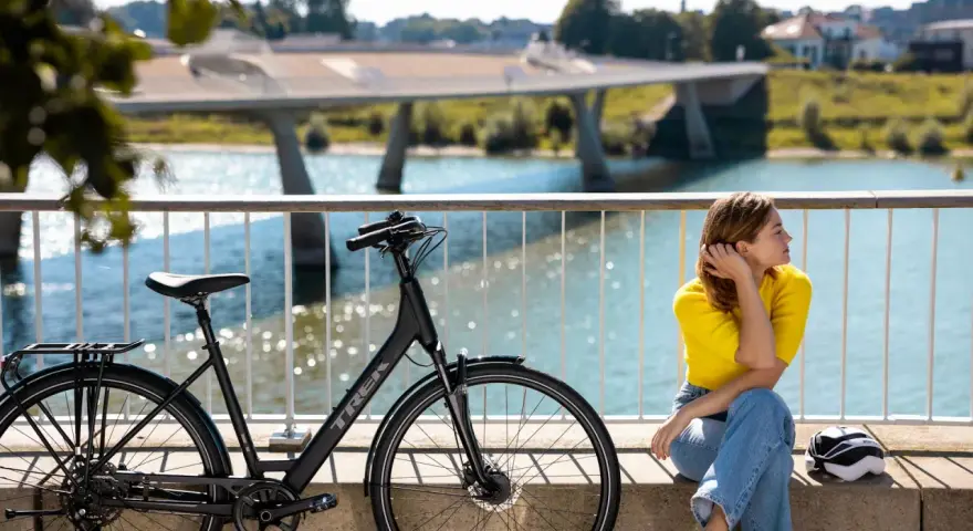 En kvinna i gula tröjan och jeans vilar vid en elcykel. Frågan "hur lång tid tar det att cykla 1 mil med elcykel" hänger i luften.