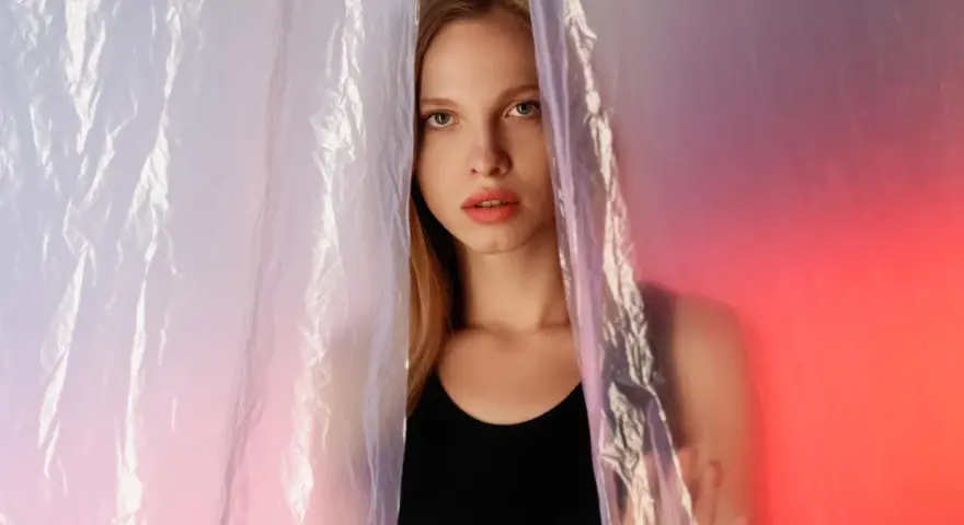 A young woman peeks through crinkled plastic sheet materials, her face illuminated by soft, colorful light.