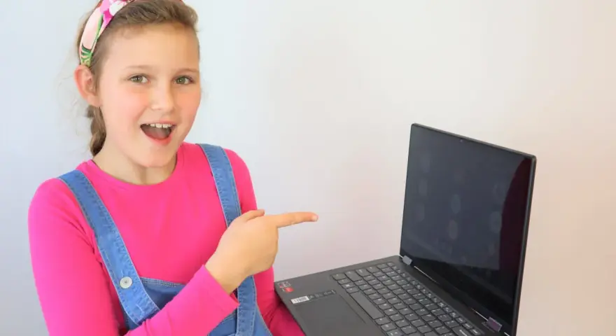 Uśmiechnięta dziewczynka w ogrodniczkach wskazuje na ekran laptopa, gdzie widać okienka z ikonami. Idealne miejsce na naukę angielskiego online dla dzieci.