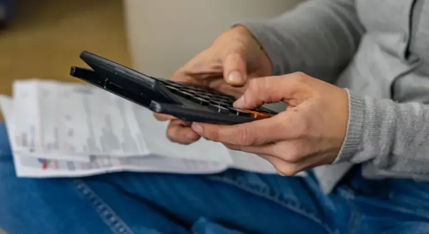 Ręce osoby w dżinsach i szarej bluzie liczą coś na kalkulatorze. Obok leżą dokumenty. Może to być rozliczenie rachunków, które zajmuje jej g12w godziny.