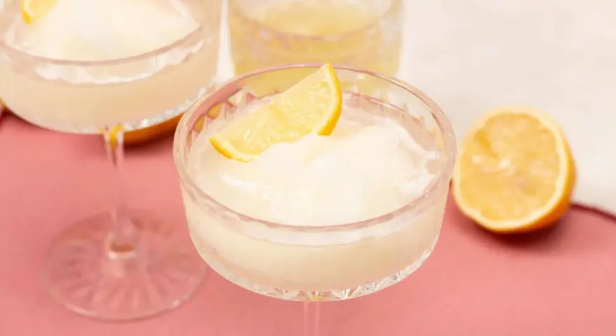 Erfrischendes Limoncello Sorbet in einem Kristallglas, garniert mit einer Zitronenspalte. Ein halbe Zitrone liegt daneben.
