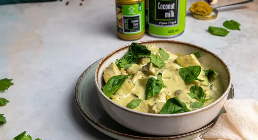Aksamitny kurczak w zielonym curry, udekorowany świeżym szpinakiem i kolendrą. Obok puszka mleka kokosowego i słoiczek pasty curry.