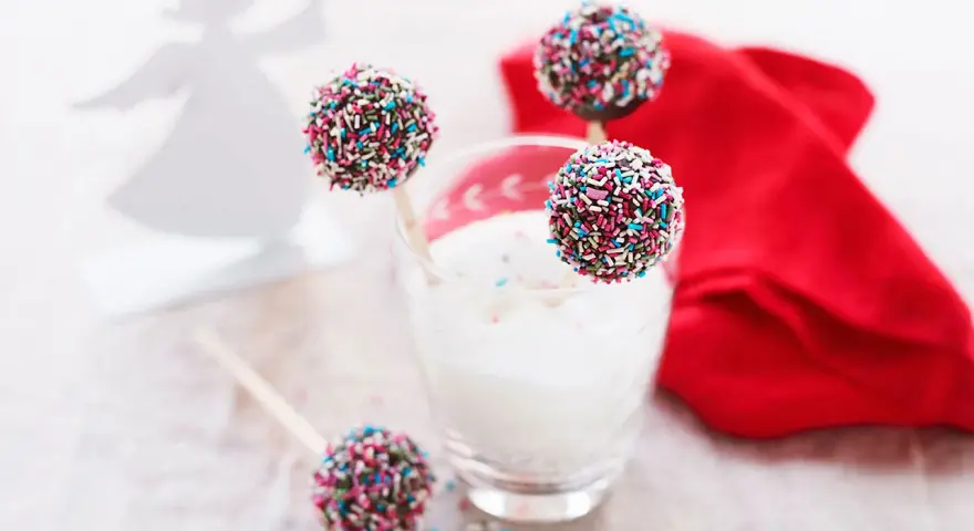 Söta cake pops med pepparkakssmak, dekorerade med färgglada strössel. En vit ängel i bakgrunden.