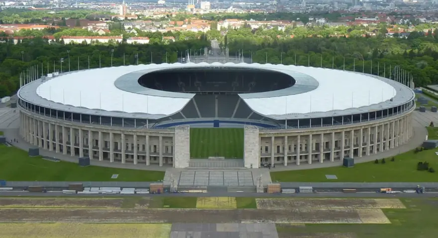 Das Olympiastadion Berlin mit seinem markanten Dach und der Skyline im Hintergrund. In der Ferne ragt der Fernsehturm, der wie ein glockenturm Berlin überragt.