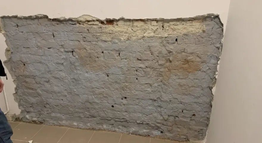 Fragment odsłoniętej, starej ceglanej ściany z szarym tynkiem i fragmentami żółtego podkładu.