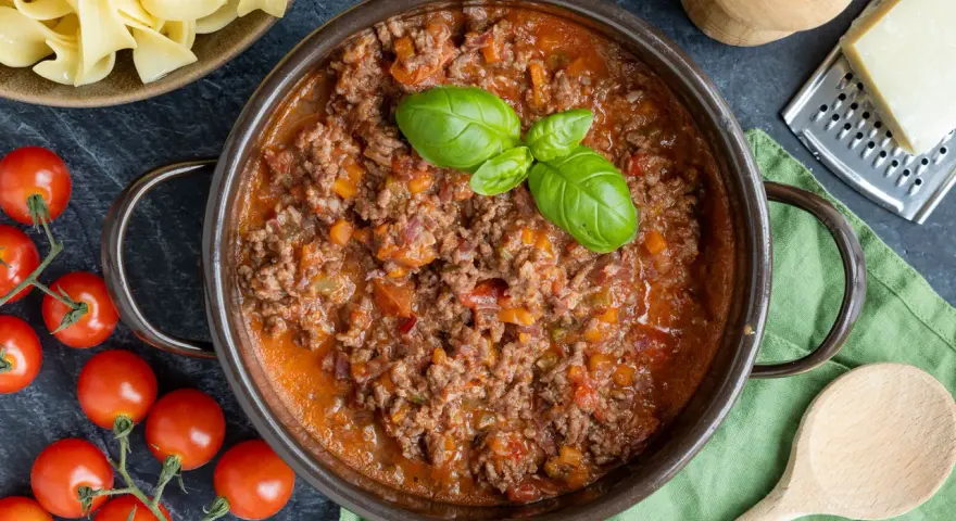 Herzhaftes Ragout Bolognese in einem Topf, garniert mit Basilikum. Daneben frische Tomaten, Bandnudeln und Parmesan.