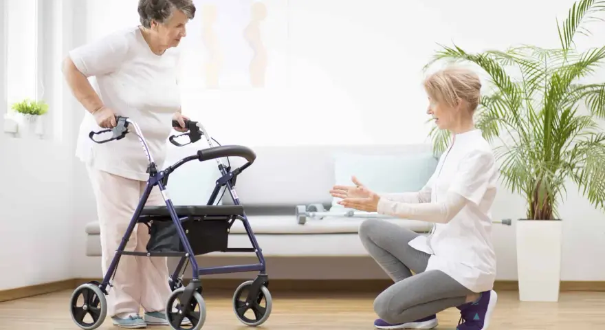 Starsza pani z pomocą rehabilitantki uczy się chodzić przy użyciu chodzika.