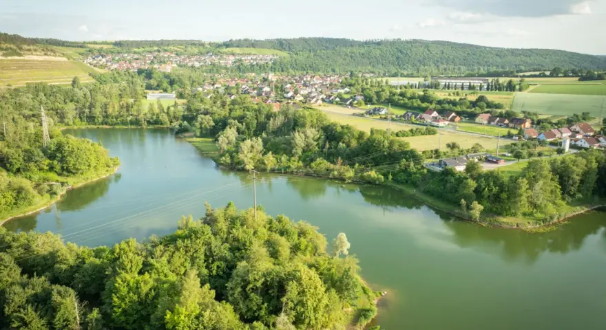 Grüne Hügel, ein Dorf und der glitzernde Klostersee Triefenstein unter blauem Himmel.