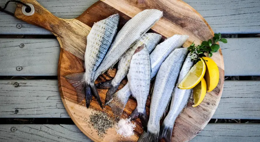 Plusieurs poissons frais sur une planche à découper en bois avec du sel, du poivre et des quartiers de citron.