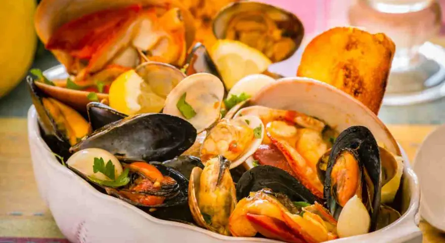 Un fumetto di pesce ricco di cozze, vongole e crostacei, guarnito con limone e prezzemolo, servito in una ciotola bianca.