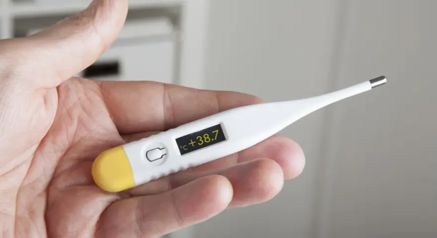 Dłoń trzyma termometr wskazujący 38.7°C. Czas na działanie, jak zbić gorączkę.