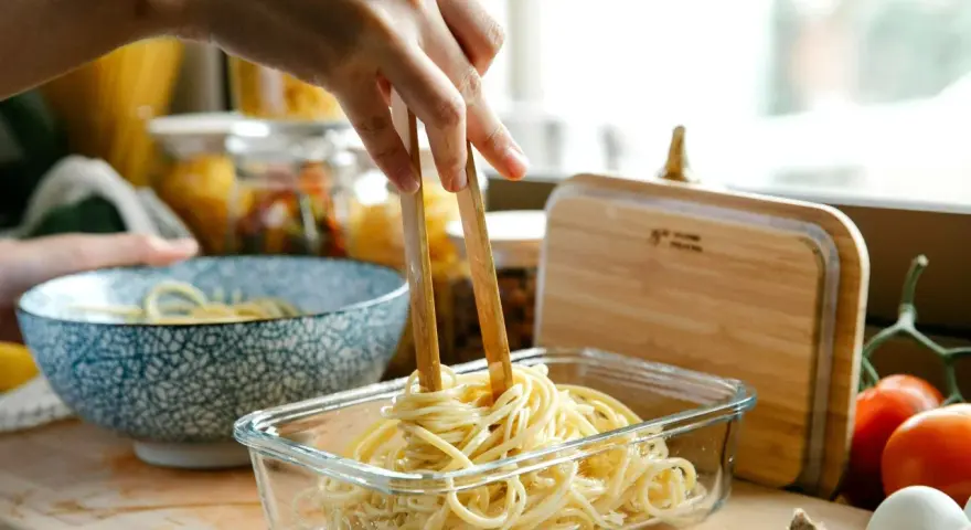 Dłoń trzymająca pałeczki wyciąga makaron spaghetti z szklanego pojemnika.