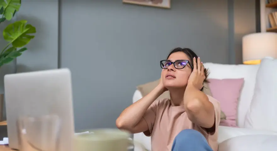 Kobieta z okularami zakrywa uszy, cierpiąc z powodu nadwrażliwości na dźwięki. W tle laptop i kanapa.