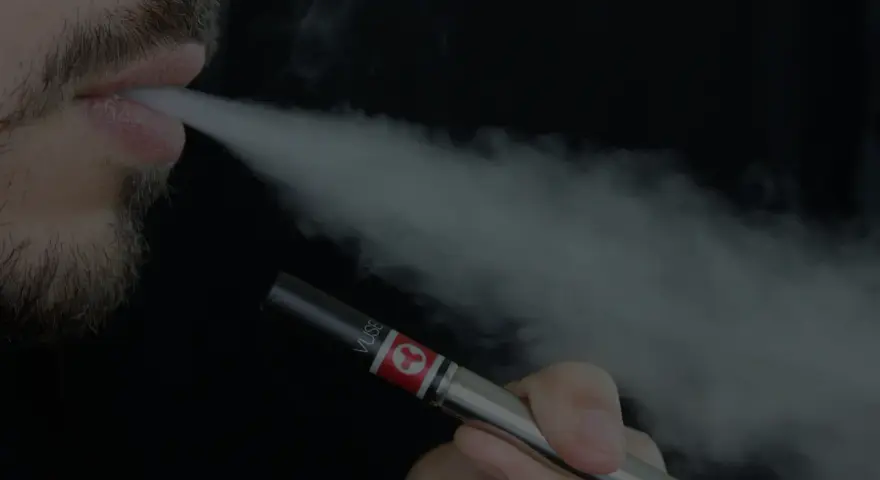 Un hombre con barba exhala vapor de un vapeador, similar al estilo Heisenberg.