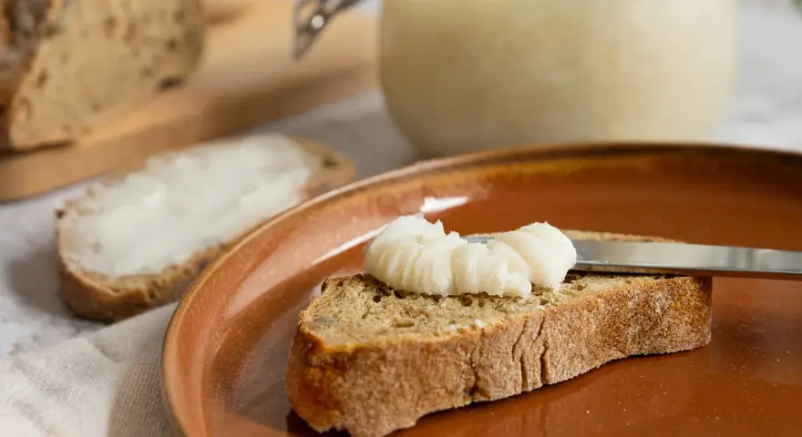 Ein Glas mit selbstgemachtem Schmalz steht bereit. Eine Scheibe Brot wird gerade mit dem Schmalz bestrichen, ein Genuss für jeden, der Schmalz selber machen möchte.