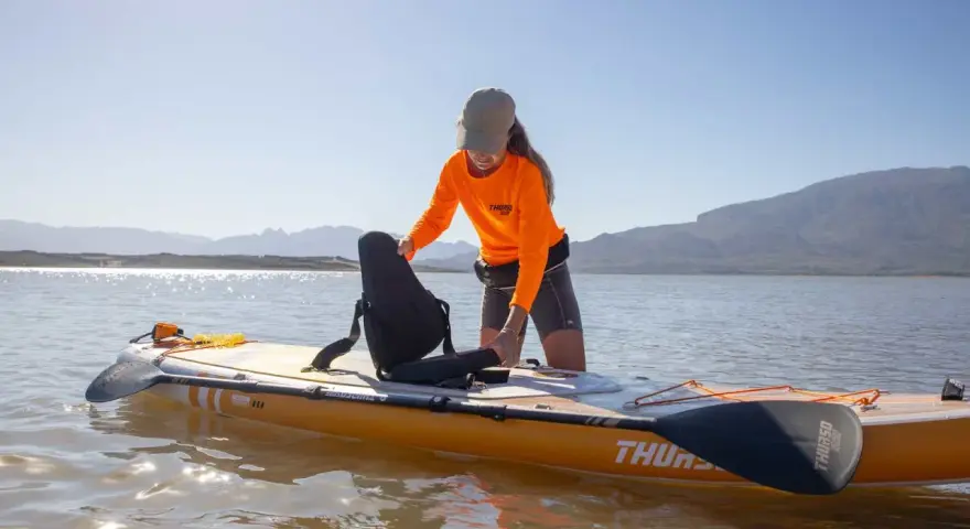 Frau richtet Sitz auf SUP-Board ein, das wie ein Kajak genutzt werden kann. Sonnenschein, Berge und See im Hintergrund.
