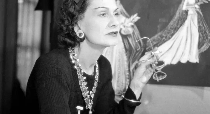 Coco Chanel, icona di stile, con occhiali in mano, sullo sfondo schizzi di abiti da sposa. Un'immagine che racconta la sua storia.