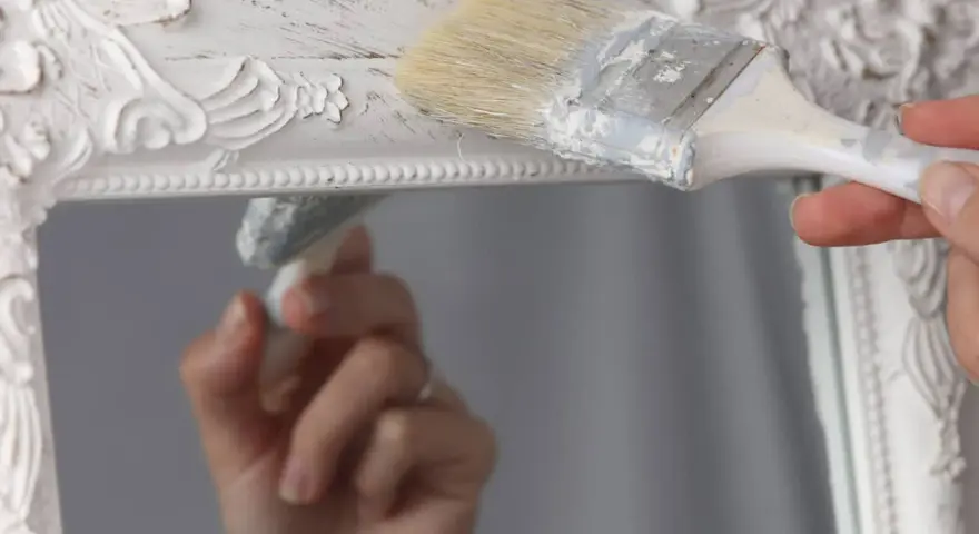 Pintura a la tiza blanco antiguo en un marco de espejo ornamentado. Una mano aplica la pintura con una brocha, creando un acabado desgastado.