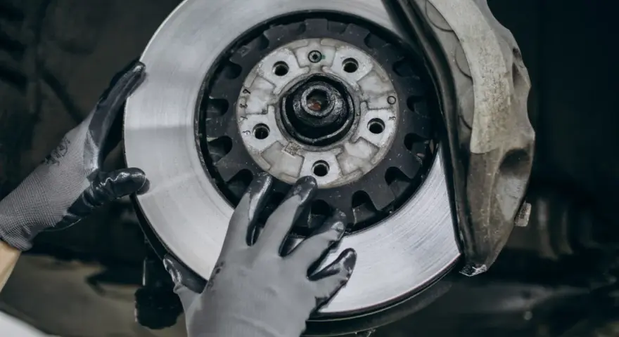 Mechanik w rękawiczkach wymienia zużyte tarcze hamulcowe. Widać nowy, lśniący element obok starego zacisku.