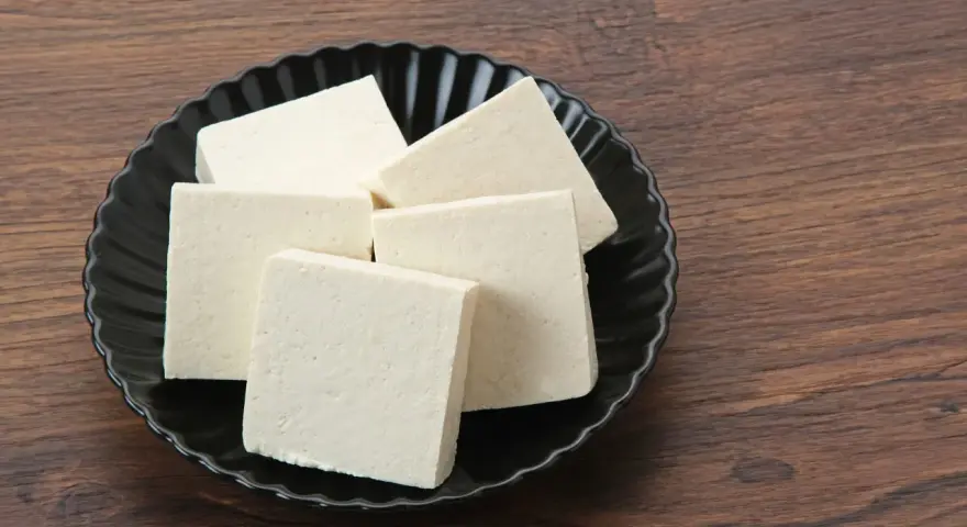 Kilka kostek białego tofu na czarnym talerzu. Czy tofu można mrozić? Tak, po rozmrożeniu zmienia teksturę, stając się bardziej gąbczaste.