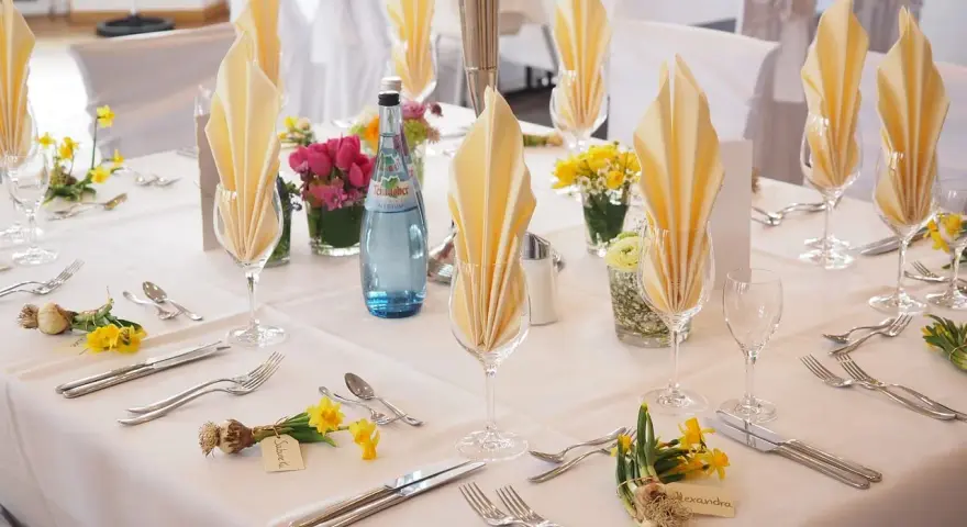 Elegancko nakryty stół weselny z żółtymi serwetkami, kwiatami i butelką wody. Każde miejsce ma imienną wizytówkę, jak dla drużby.