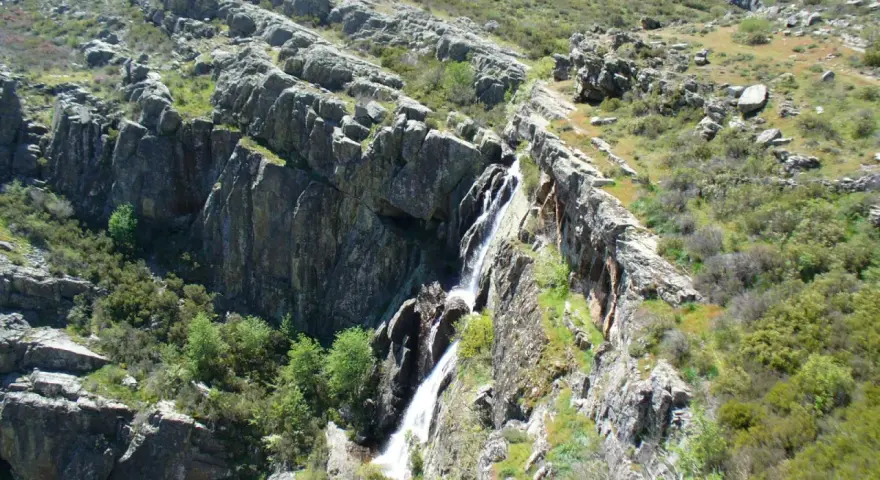 Cascada cayendo por rocas escarpadas cerca de un pico ocejon, rodeado de vegetación verde y rocas.